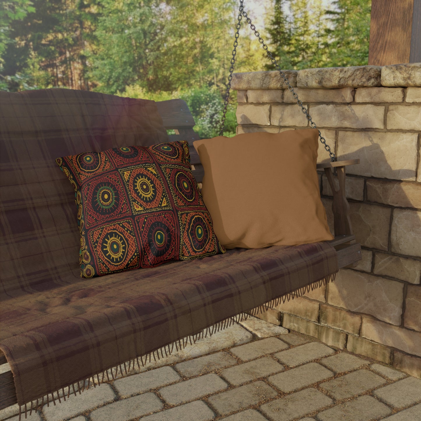 A wooden swing bench with a brown plaid blanket and two cushions—one solid tan and one with an African-inspired geometric pattern—sits on a stone patio by a stone wall, with green trees visible in the background.