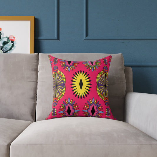 A vibrant throw pillow with bold, geometric, and symmetrical sunburst patterns in yellow, black, green, and purple on a bright pink background adds colorful home decor flair to a light gray sofa beneath a blue wall. A framed cactus print is partially visible on the left.