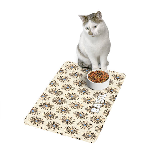 A white and gray cat sits behind a food bowl filled with kibble, placed on a beige mat featuring an African-inspired geometric starburst pattern and two cartoon cats. The background is plain white.