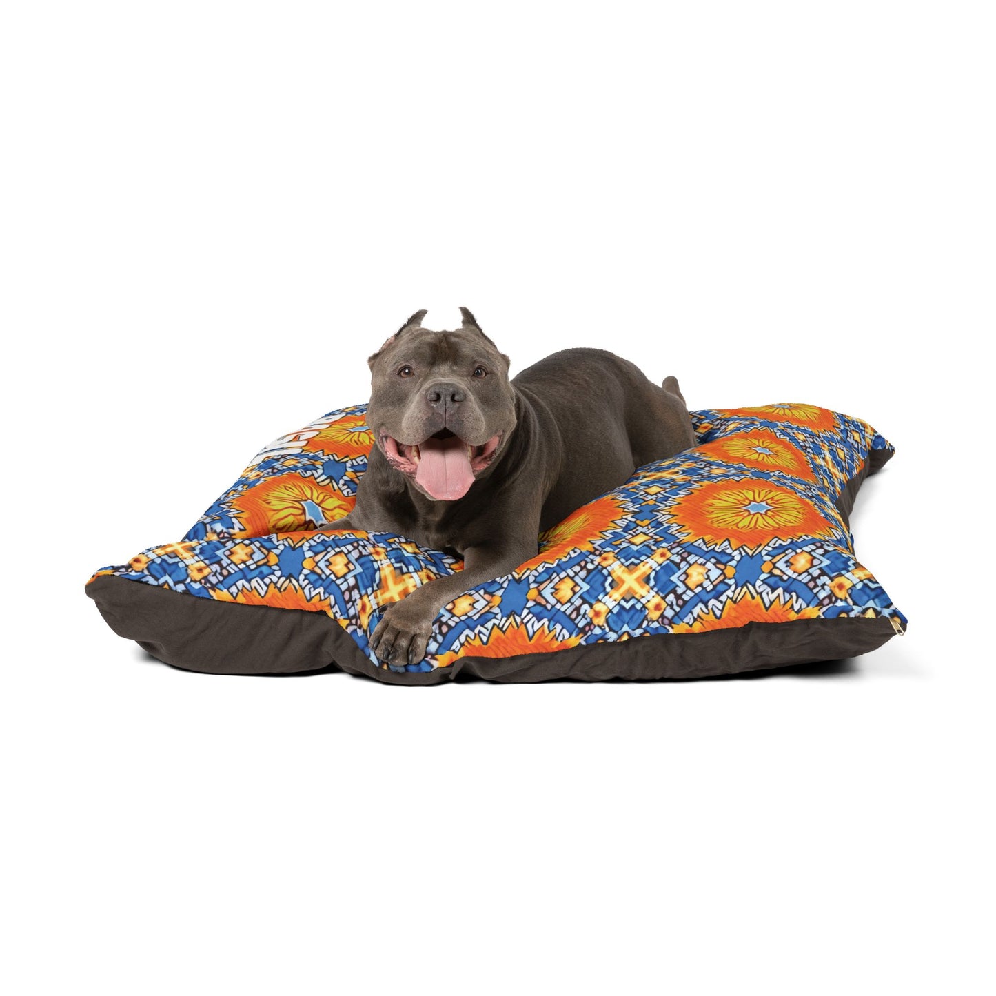 A gray pit bull with cropped ears lies on a stylish dog bed featuring vibrant blue, orange, and yellow geometric designs. The dog looks happy with its tongue out and front legs extended, relaxing on this eye-catching and comfy accessory against a plain white background.