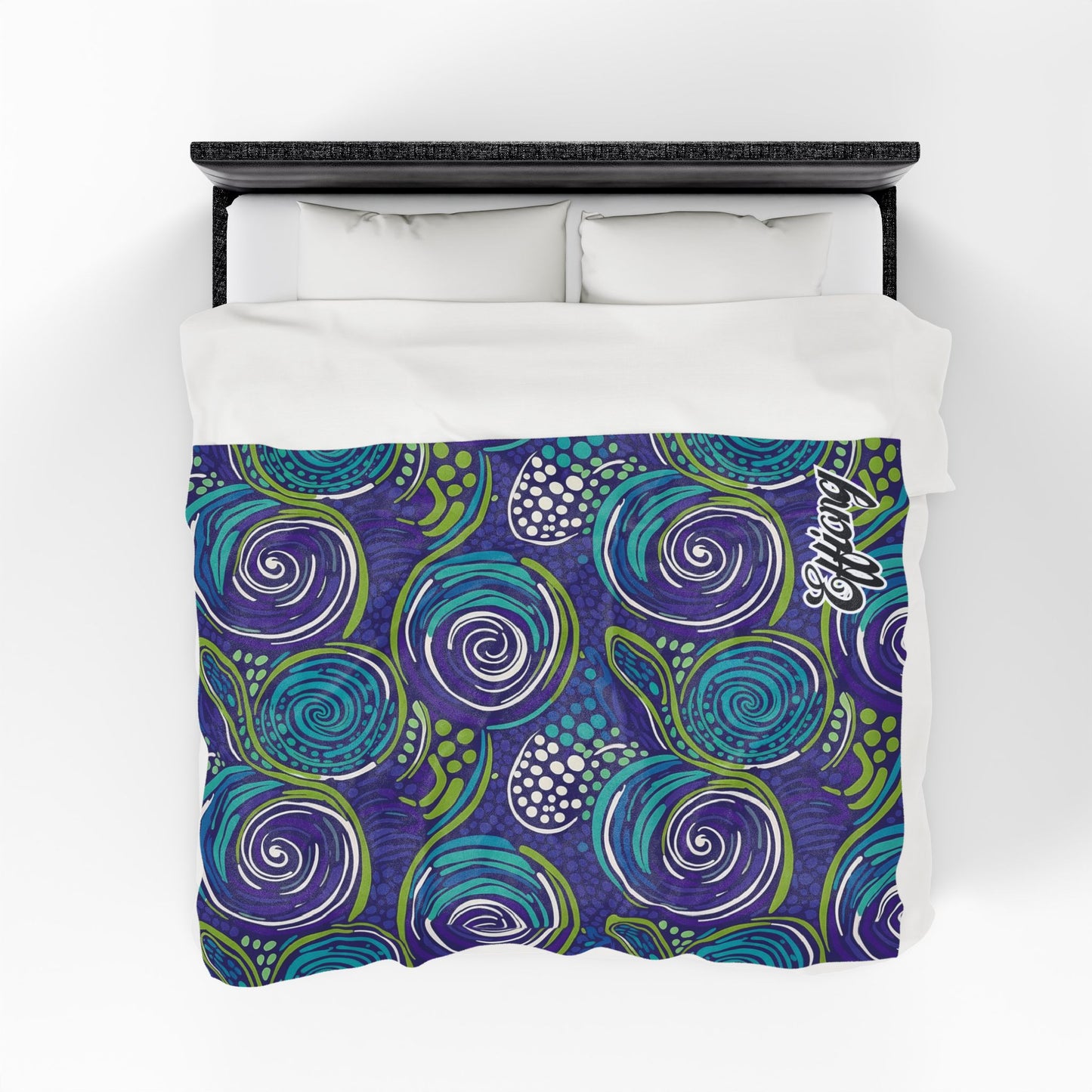 A bed with a white pillow and sheet set is topped by a personalized velveteen blanket featuring a vibrant, abstract swirl pattern in blue, purple, and green with white dotted accents and the Coastal logo. The photo is taken from an overhead view.