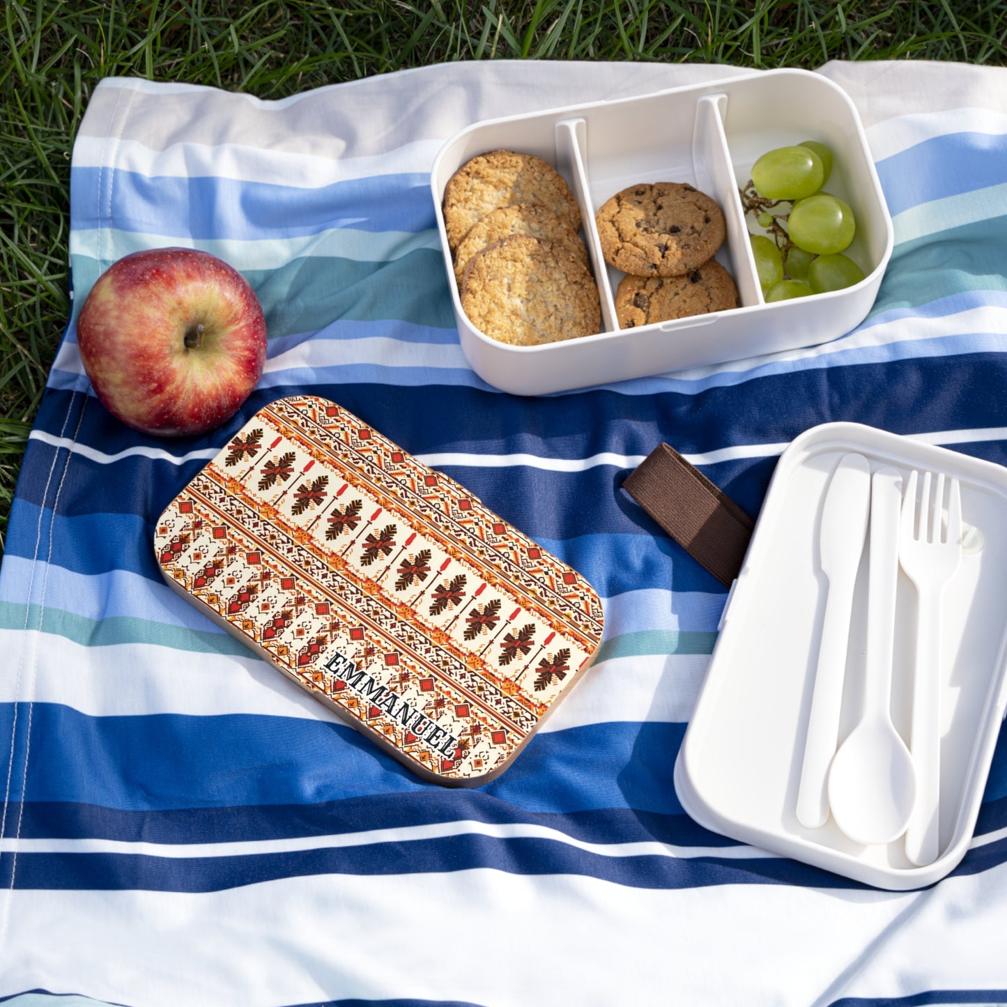 A personalized lunch box with a patterned lid labeled “Emmanuel” sits open on a blue-striped picnic blanket. Inside are cookies and green grapes. An apple, white reusable cutlery, and green grass complete the cheerful scene.