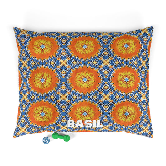 Rectangular personalized pet bed with a vibrant orange, yellow, and blue geometric lion face pattern. The name BASIL appears in bold white letters near the bottom. In front are two dog toys: a blue ball and a green bone-shaped chew toy.
