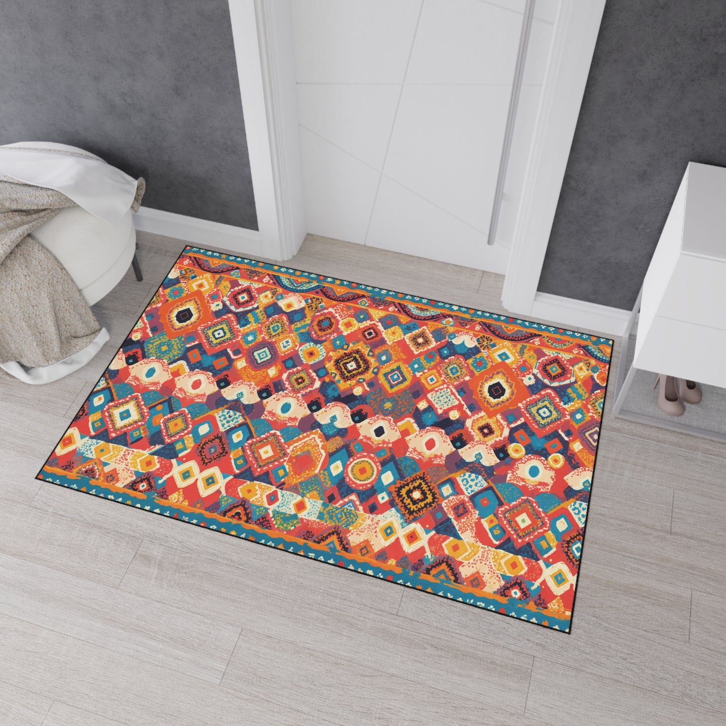 A colorful rectangular rug with African-inspired aesthetics and geometric and floral patterns in red, blue, yellow, and orange lies on a light wood floor near a white door, gray walls, a white cabinet, and a beige throw blanket draped on the left side.