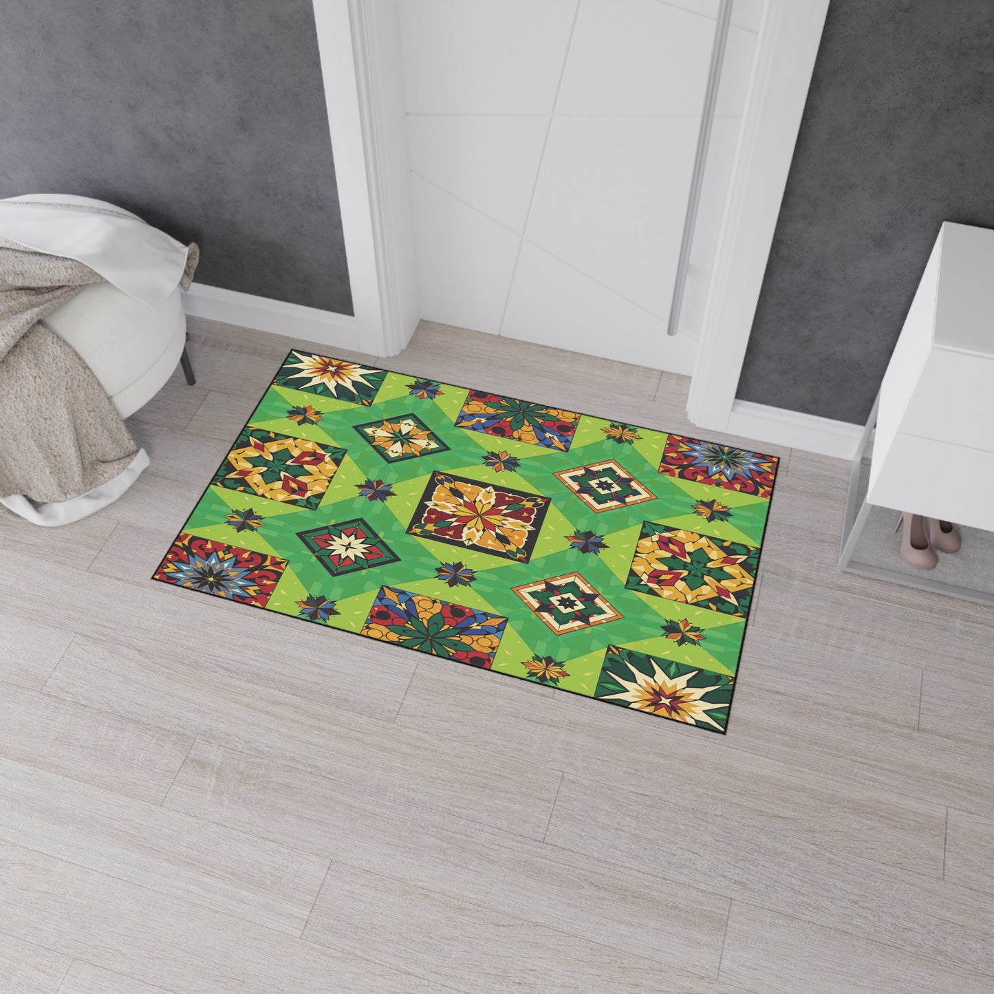 A colorful rectangular rug with a green geometric patchwork pattern, floral motifs, and African-inspired aesthetics lies on light wood flooring in front of a white door, near a gray wall, a white dresser, and a beige throw blanket draped over a chair.
