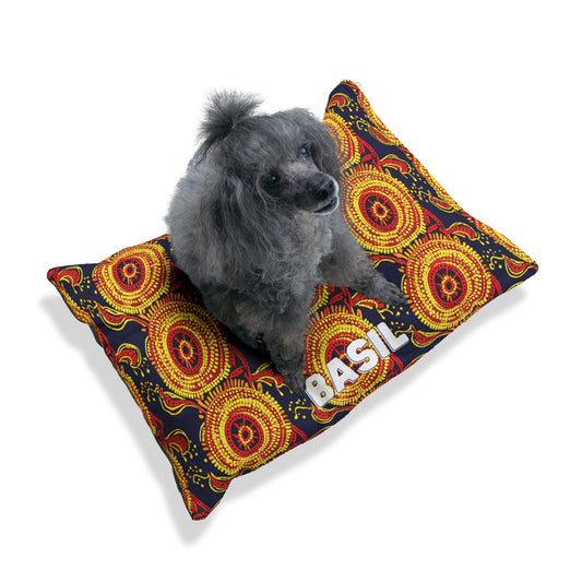 A gray poodle with curly fur sits on a rectangular dog bed featuring an African-inspired pattern of vibrant red, yellow, and orange circles. The bed has the name “BASIL” in large white letters. The background is plain white.