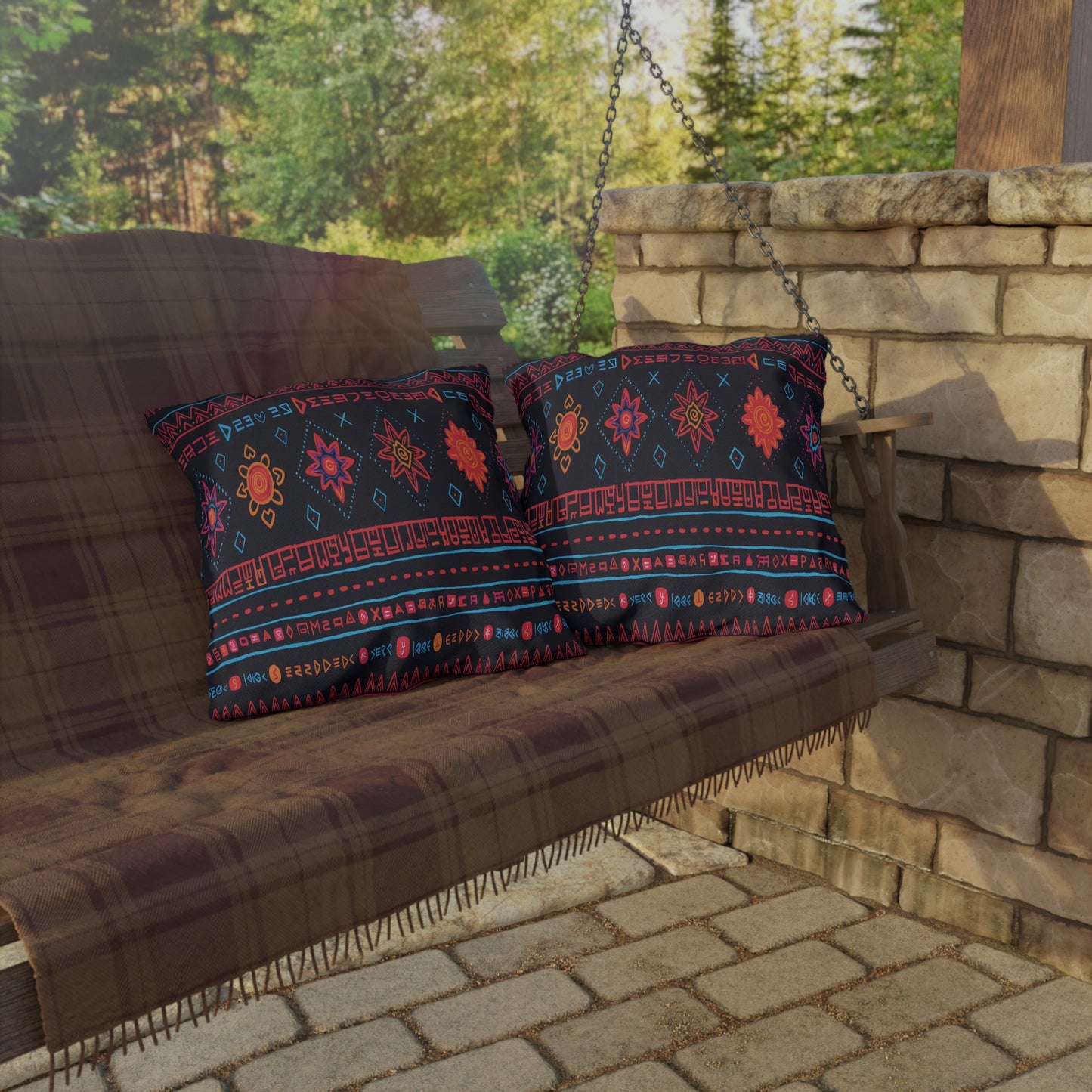 A wooden porch swing with an African-inspired brown plaid throw blanket, two colorful patterned pillows, and black metal chains hangs near a stone wall. Sunlight filters through trees, casting reflections and shadows on the patio’s brick floor.
