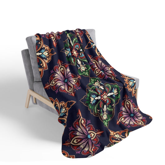 A soft, navy blue sherpa fleece blanket with vibrant floral and paisley patterns in red, orange, purple, green, and beige drapes over a modern gray armchair with wooden legs, adding cozy home decor flair against a white background.