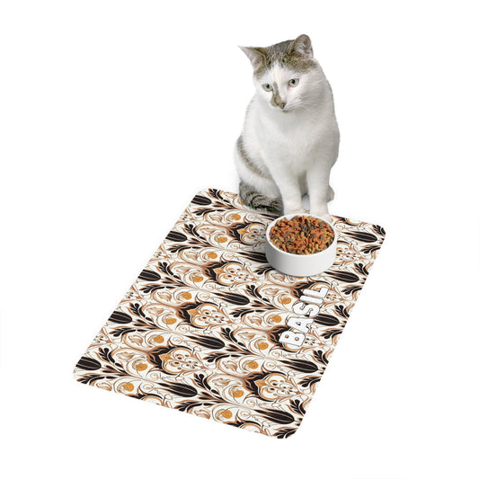 A white and gray cat sits on an African-inspired mat with brown, tan, and black swirls. A white bowl of dry cat food rests on the mat, which has the word BASIL printed in block letters. The background is white.