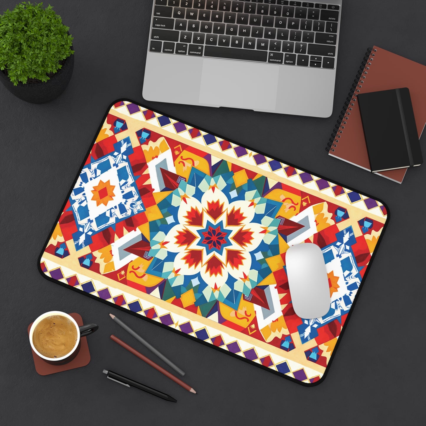 A colorful workspace accessory, this neoprene desk mat features a bold geometric and floral pattern in red, blue, yellow, and teal on a black desk alongside a mouse, laptop, notebooks, plant, pen, pencil, and coffee.