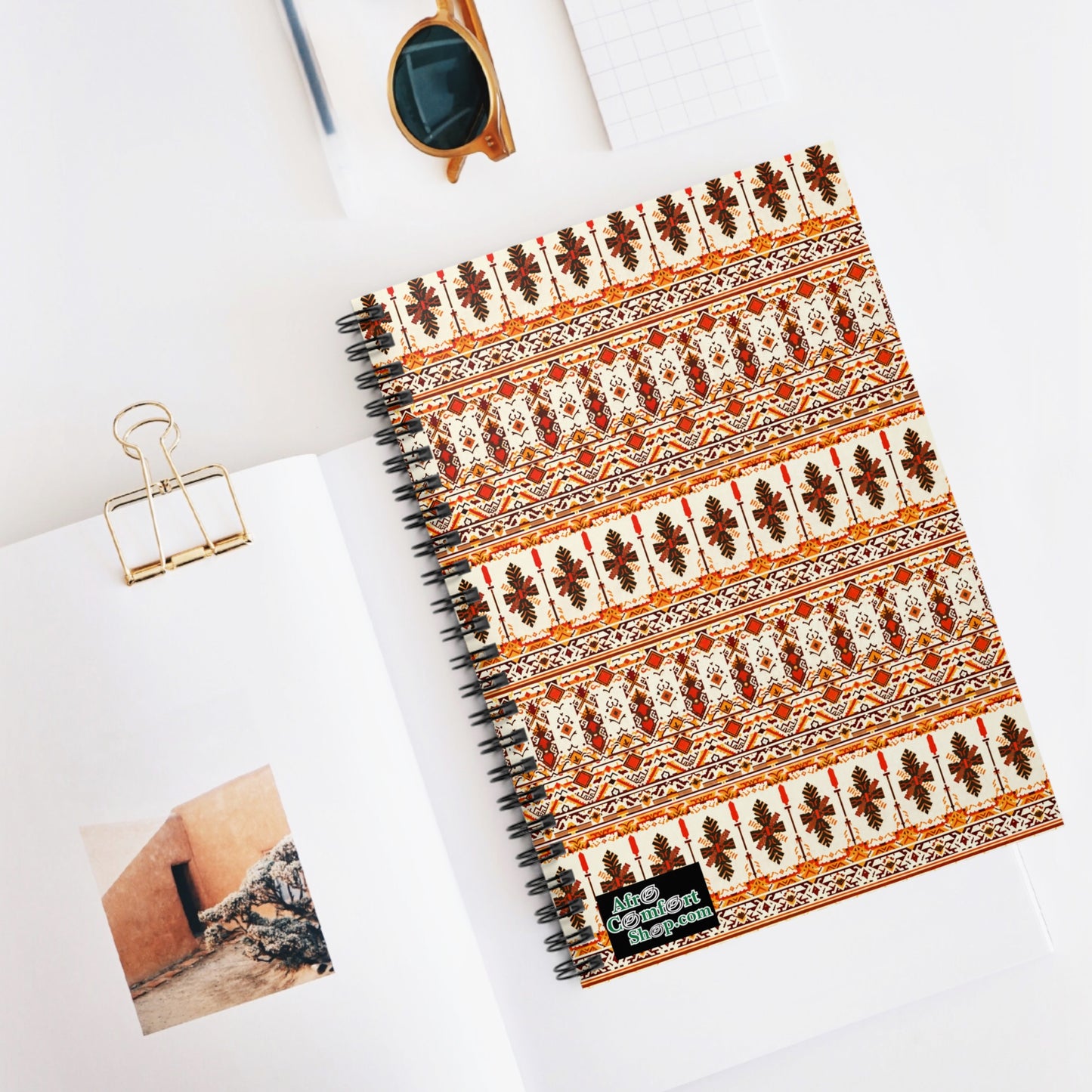 A premium writing notebook with a floral and geometric cover, inspired by African design, lies on an open book showing a building photo. Nearby are brown sunglasses, a gold binder clip, and graph paper on a clean white surface.