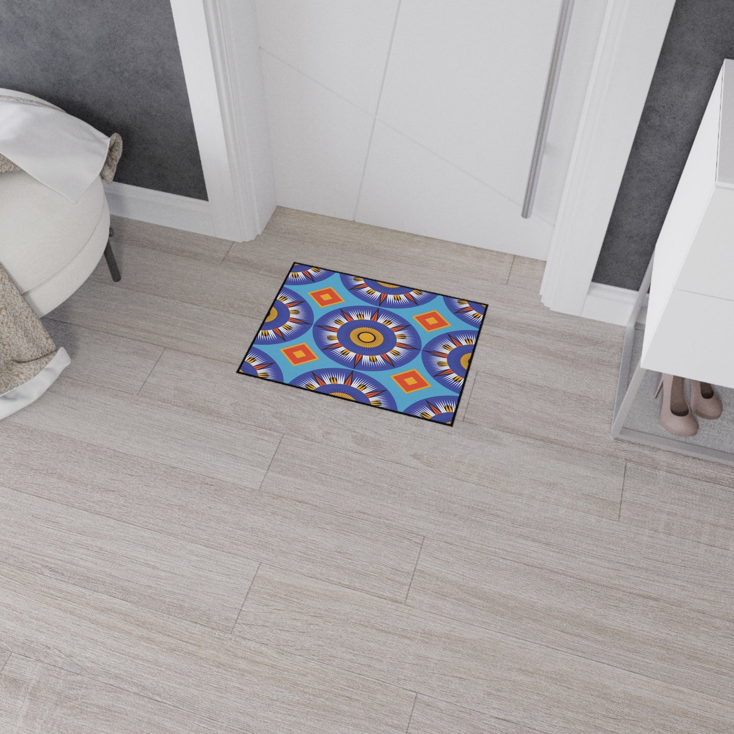 A small, rectangular mat with a vibrant geometric mandala pattern in blue, yellow, orange, and purple lies on light wood flooring near a white door. Its non-slip rubber backing keeps it secure next to a white dresser holding beige heels and a white chair.