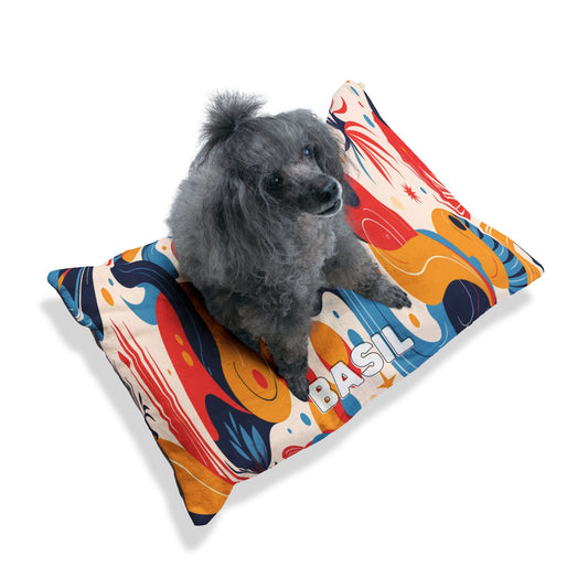 A small gray poodle with curly fur sits on a colorful, African-inspired abstract-patterned pillow. The pillow features swirls of red, blue, gold, and white, and has the name BASIL printed in bold capital letters on the side. The background is plain white.