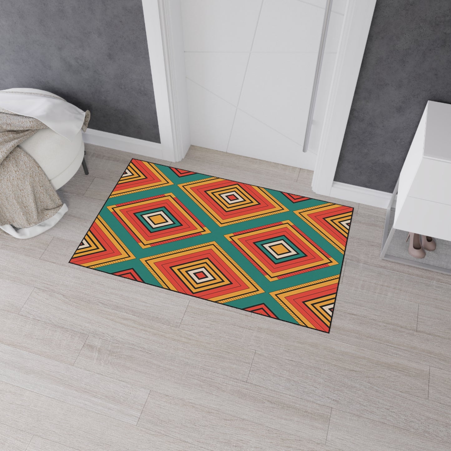 A rectangular African-inspired doormat with a bold geometric diamond pattern in red, yellow, orange, white, and teal sits on light wood flooring by a door. It features non-slip rubber backing for added safety near a white cabinet and beige throw-draped chair.