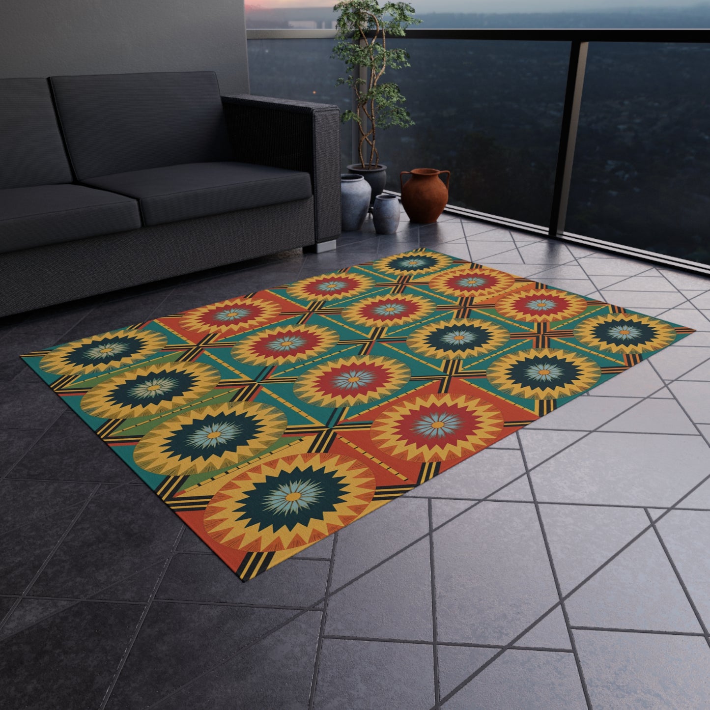 A geometric, multicolored rug in African-inspired outdoor style with circular starburst patterns in yellow, red, green, and blue lies on a tiled balcony floor beside a dark gray sofa. Potted plants and a railing overlook a nighttime cityscape.