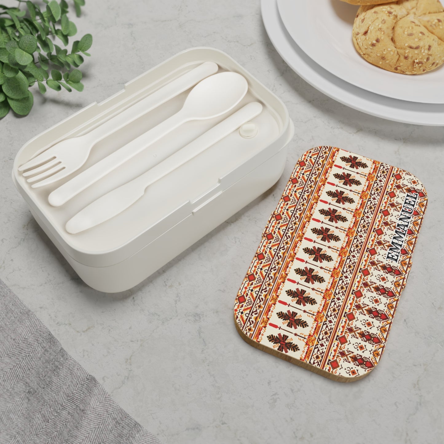 An African-inspired bento lunch box with a lid featuring an ornate red and brown geometric pattern and the word EMMANUELLE sits on a marble surface beside cookies, greenery, and cutlery—perfectly BPA-free for your personalized meals.
