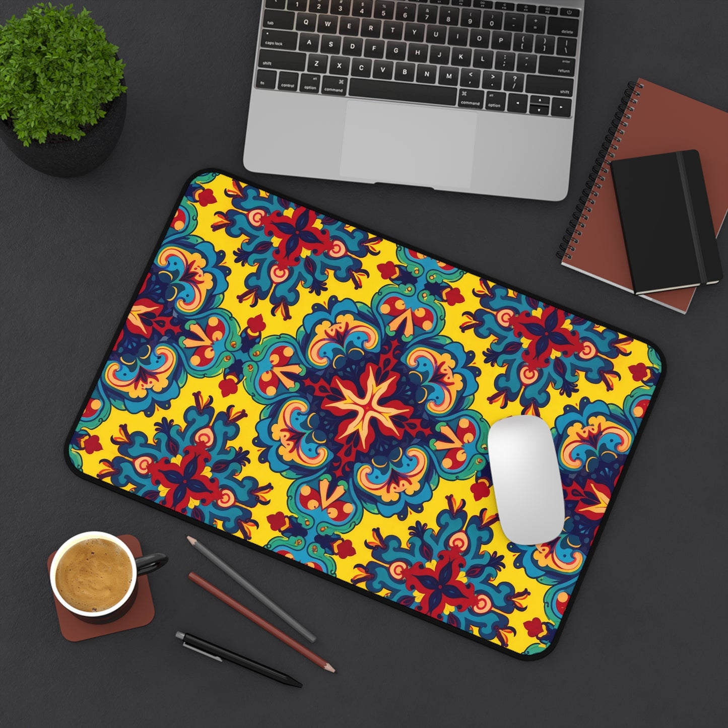 A colorful neoprene desk mat with a bold, symmetrical floral pattern in blue, red, and yellow brightens up a dark desk, next to a laptop, brown notebooks, a potted plant, coffee cup, two pencils, and a white mouse.