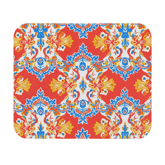 A rectangular mat with rounded corners featuring a vibrant, ornate pattern. The background is bright red with symmetrical yellow, blue, and white floral and leaf designs—an eye-catching African-inspired mouse pad with a non-slip rubber base.