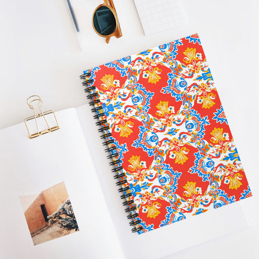 A premium writing notebook with vibrant red, blue, yellow, and white floral and geometric patterns lies on a white desk beside an open book, gold binder clip, brown sunglasses, and a small checkered notepad.
