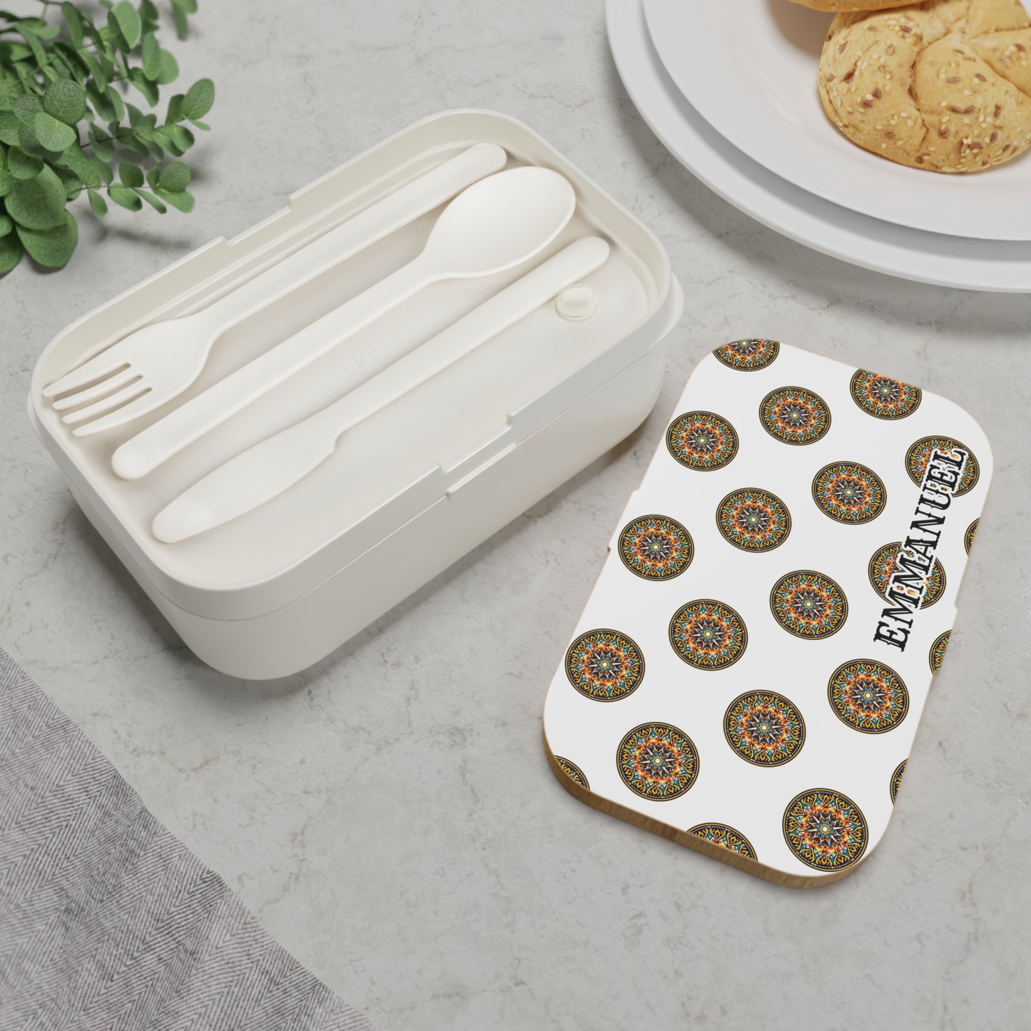 A personalized, BPA-free lunchbox with a colorful mandala lid and EMMANUEL in bold text sits on marble. This African-inspired bento lunch box includes a built-in compartment for utensils, with plates of cookies arranged nearby.