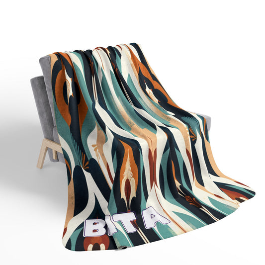 A modern, African-inspired fleece blanket with an abstract pattern in earth tones drapes over a light grey chair. The bold letters BITA decorate the bottom edge of this cozy sherpa fleece. The background is plain white.