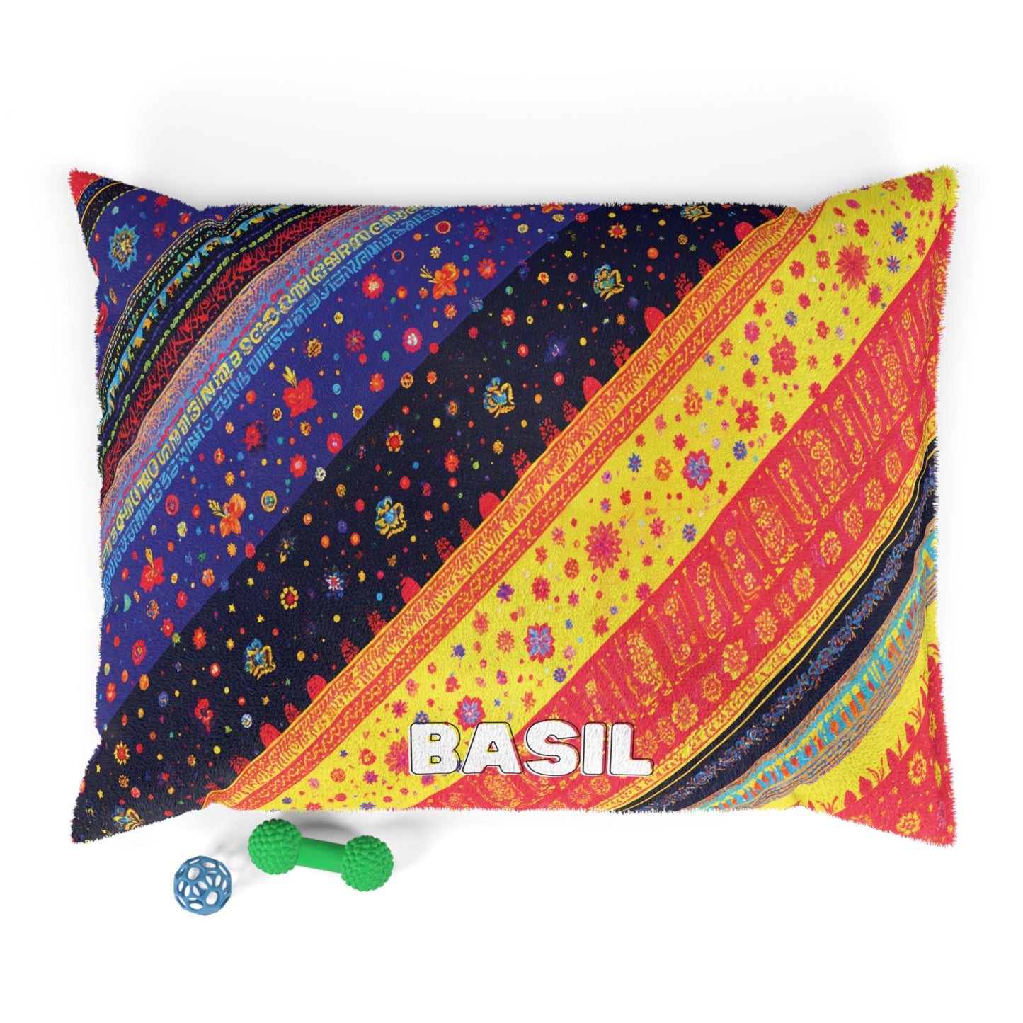 A colorful, African-inspired rectangular pillow with diagonal vibrant patterns in blue, red, yellow, and orange features the word BASIL in white letters. Two dog toys—a green bone and a blue ball—lie beside it on a white background.