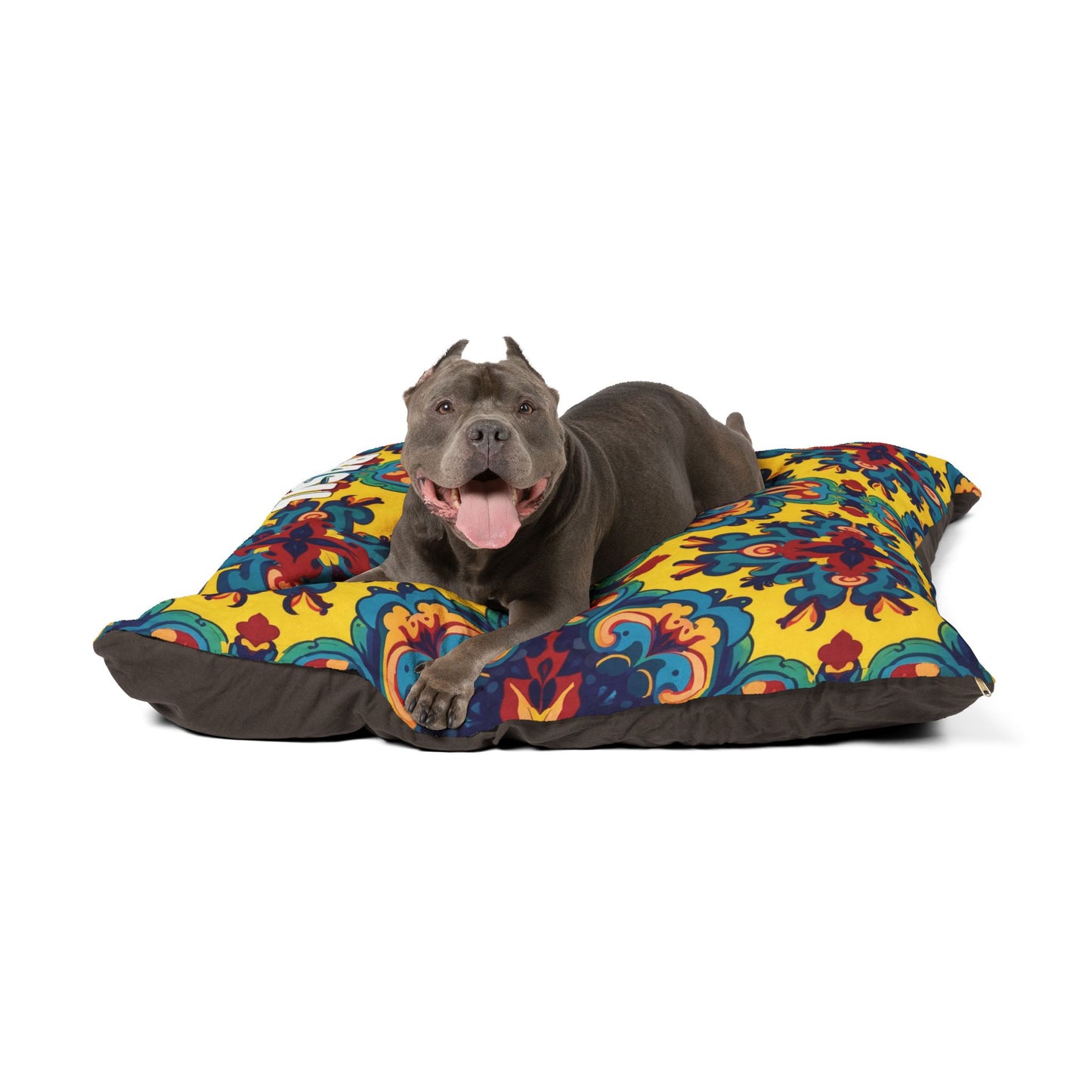 A happy gray pit bull dog lies on a stylish pet bed with a vibrant, floral pattern in yellow, blue, red, and green. Panting and tongue out, it looks at the camera with front paws stretched forward against a plain white background.