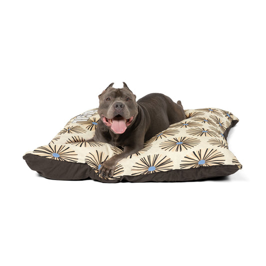 A gray pit bull with cropped ears lies on a large, rectangular dog bed featuring an African-inspired beige and brown starburst pattern, looking forward with its mouth open and tongue out, appearing happy and relaxed. The background is plain white.