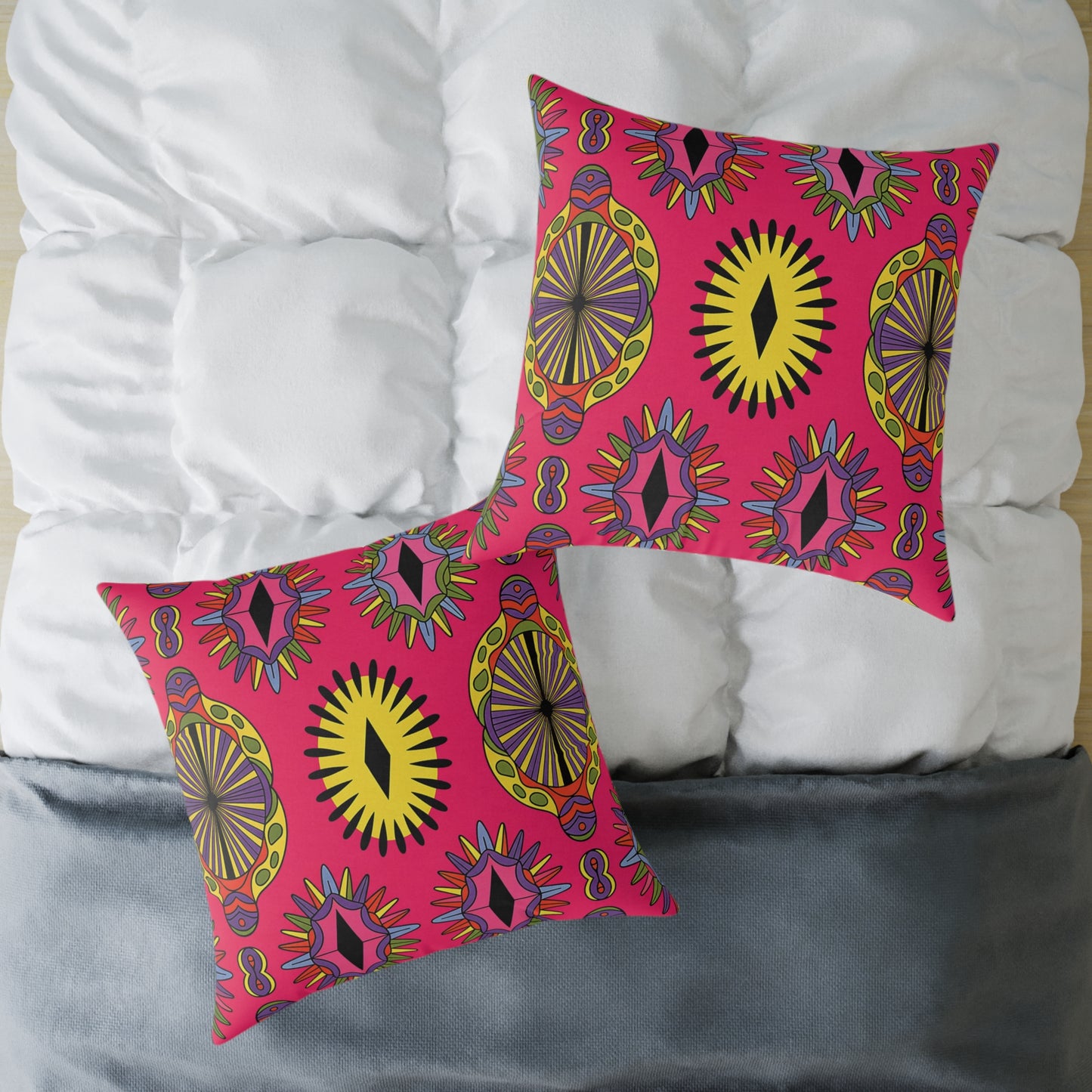 Two square pillows with bright, African-inspired geometric patterns in yellow, black, orange, and purple on a pink background rest on a white, quilted bed comforter with a gray bottom edge. These vibrant accent pillows are slightly overlapping and diagonally placed.