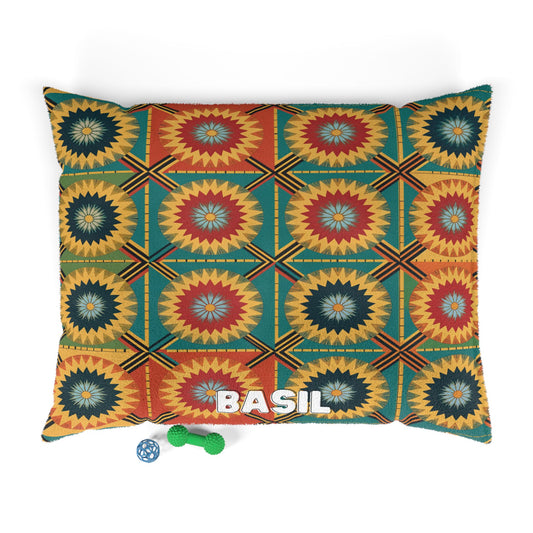 A rectangular personalized pet bed with a bold, geometric starburst pattern in green, yellow, red, and orange. “BASIL” is embroidered in white letters at the bottom. Durable construction and two dog toys complete the cozy setup.