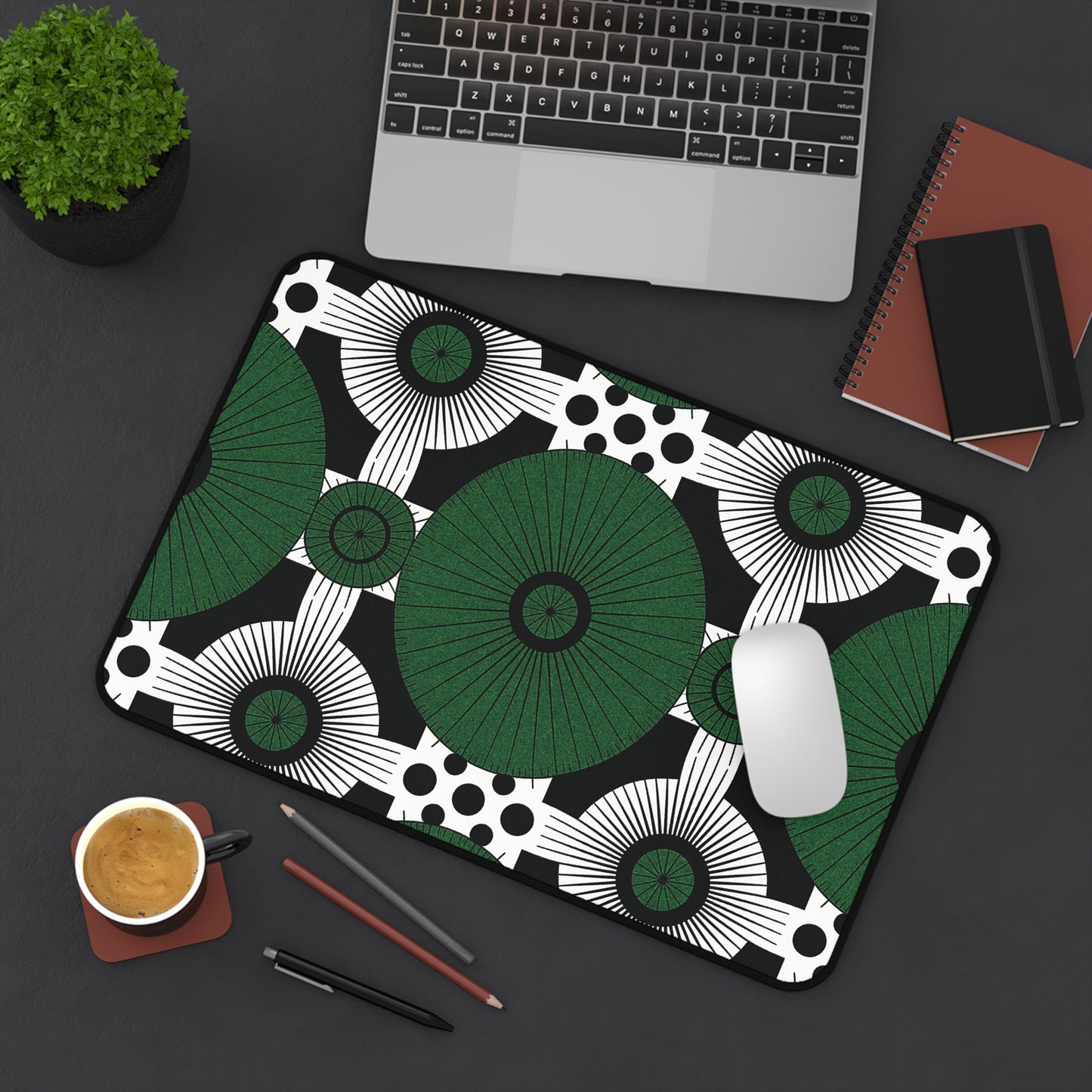 A large neoprene desk mat with a bold black, white, and green geometric pattern sits on a dark desk. Nearby are a laptop, notebooks, a smartphone, pens, coffee, and a potted plant. A white mouse rests on this colorful workspace accessory.