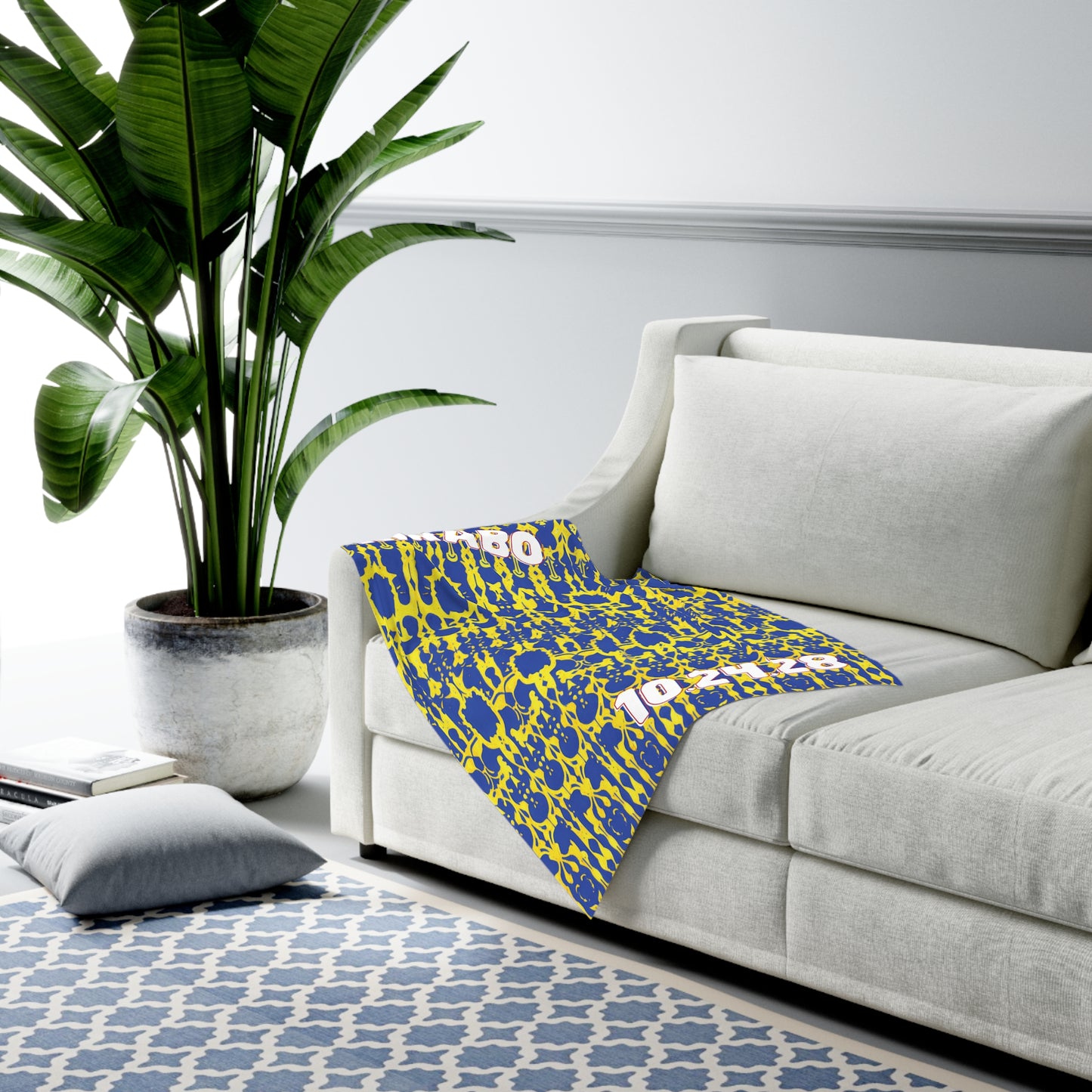 A modern living room with a large potted plant next to a white sofa. A blue and yellow African-inspired decor throw blanket is draped over the sofa. A light blue geometric rug and gray floor cushion rest on the light gray walls with darker molding.