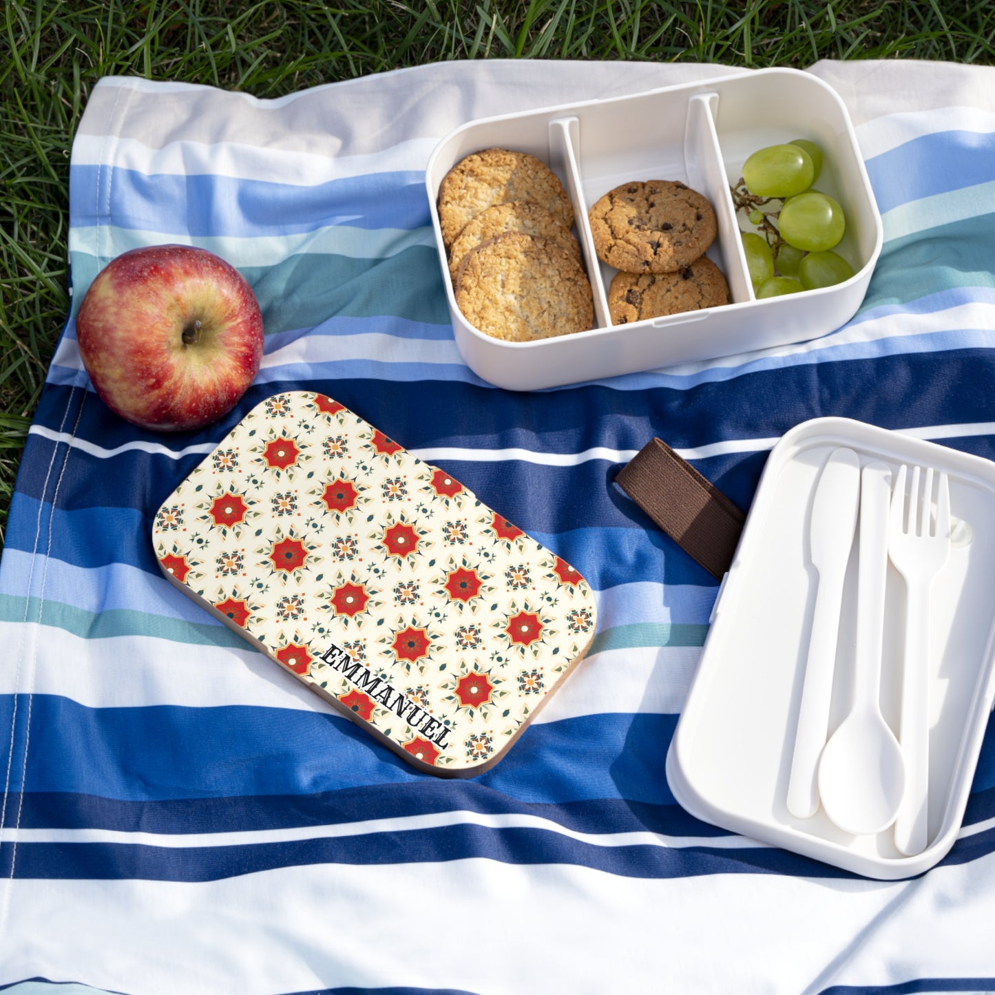 A picnic scene on grass features a striped blue-and-white blanket with an apple, grapes, cookies in a personalized lunch box, a floral lunchbox lid labeled EMMANUEL, and a white container holding a fork, spoon, and knife.
