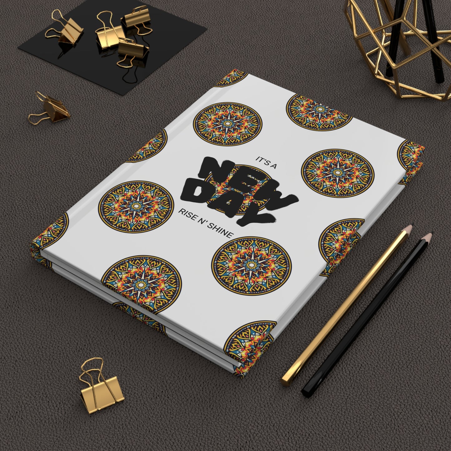A white wellness journal featuring colorful mandala patterns and the words ITS A NEW DAY RISE N SHINE lies on a textured dark surface. Gold and black pencils, binder clips, and geometric gold decor complete the look.