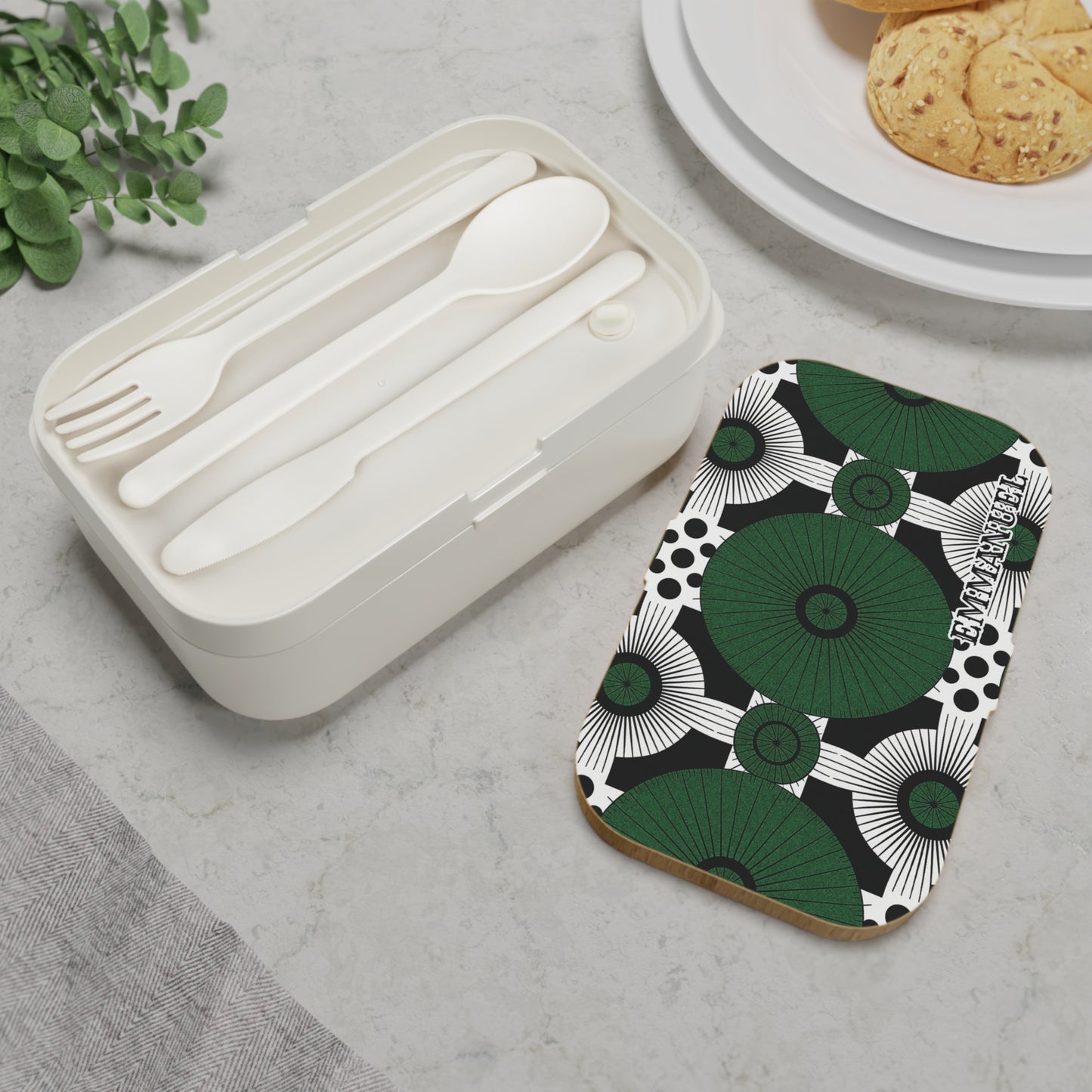 A white personalized bento lunch box with a matching spoon, fork, and knife sits on a marble surface next to a rectangular item featuring a bold green, black, and white abstract pattern. Part of a plate with cookies and greenery are also visible.