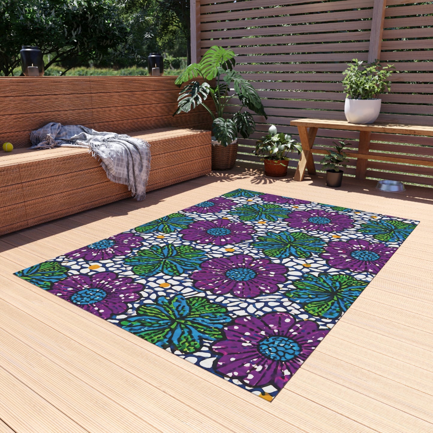 A modern patio with light wood flooring features an African-inspired outdoor rug with bold purple flowers and green leaves. A wicker sofa with a gray throw, potted plants, a wooden bench, and a privacy screen complete the serene outdoor setting.