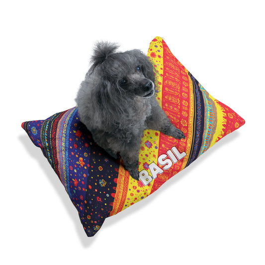 A small gray poodle sits on a colorful, patterned rectangular pillow with African-inspired red, yellow, blue, and black designs. The name “BASIL” appears in large white letters against a plain white background.