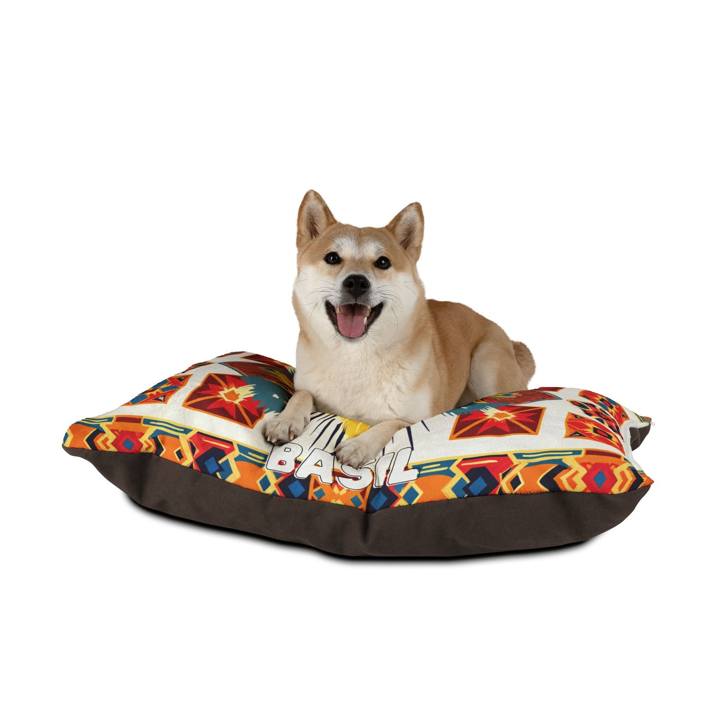 A Shiba Inu dog with tan and white fur lies on a colorful, African-inspired patterned dog bed with a dark brown base, looking at the camera with its mouth open and tongue out, appearing happy. The bed has geometric and southwestern designs. The background is white.