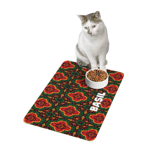 A white and gray cat sits on a vibrant, African-inspired mat with red and orange floral patterns and the word “BASIL” printed in white. Facing forward, the cat has a round bowl of dry cat food on the mat before it. The background is plain white.
