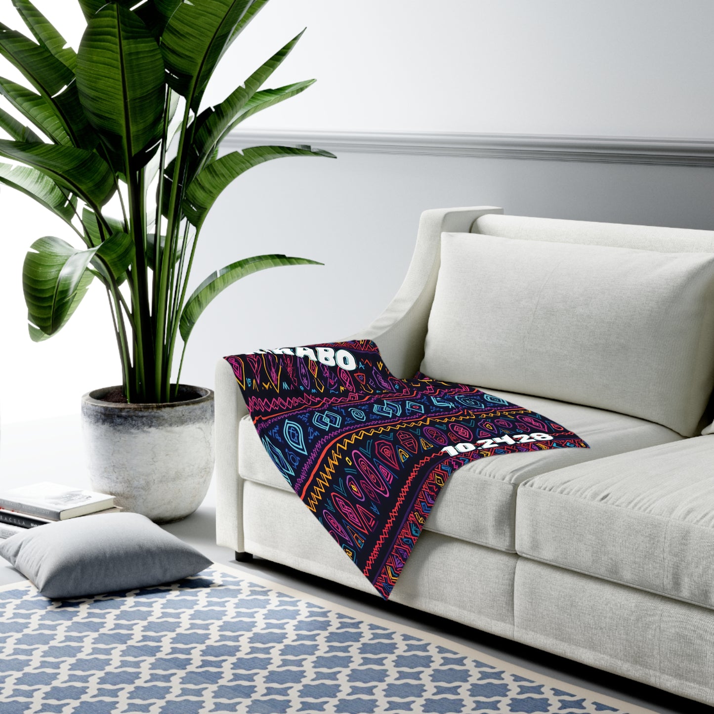 A white sofa with a colorful patterned swaddle blanket draped over one arm sits next to a large potted plant. A blue-and-white geometric rug covers the floor, and a gray floor cushion rests nearby in this modern, African-inspired living room.