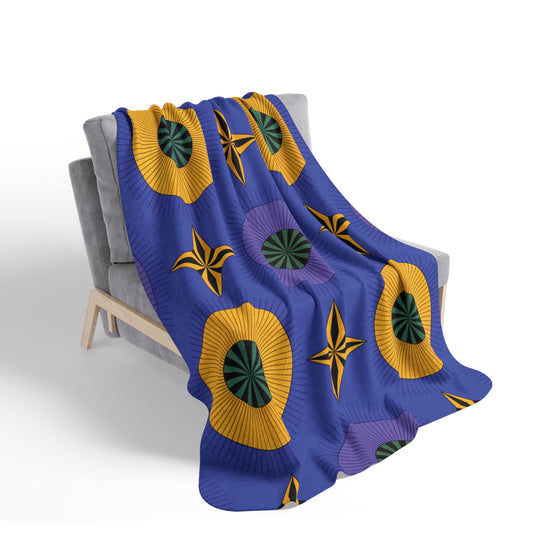 A gray, modern-style armchair with wooden legs is draped with an African-inspired fleece blanket. The blanket features a blue background with large yellow and purple geometric flower patterns, green and black centers, and yellow star shapes.