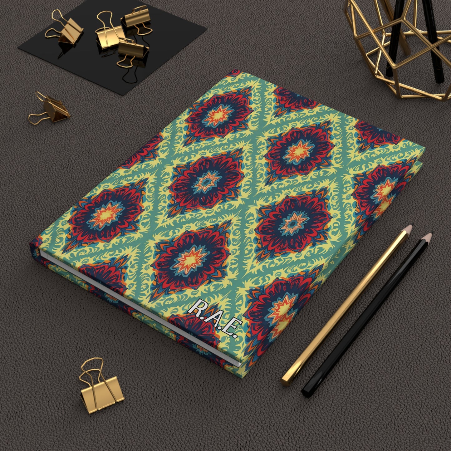 A vibrant African-inspired personalized journal with a colorful geometric floral pattern lies on a textured brown surface. Nearby are gold binder clips, black and gold pencils, and a gold wire decor piece. The notebook’s cover reads PLACE.
