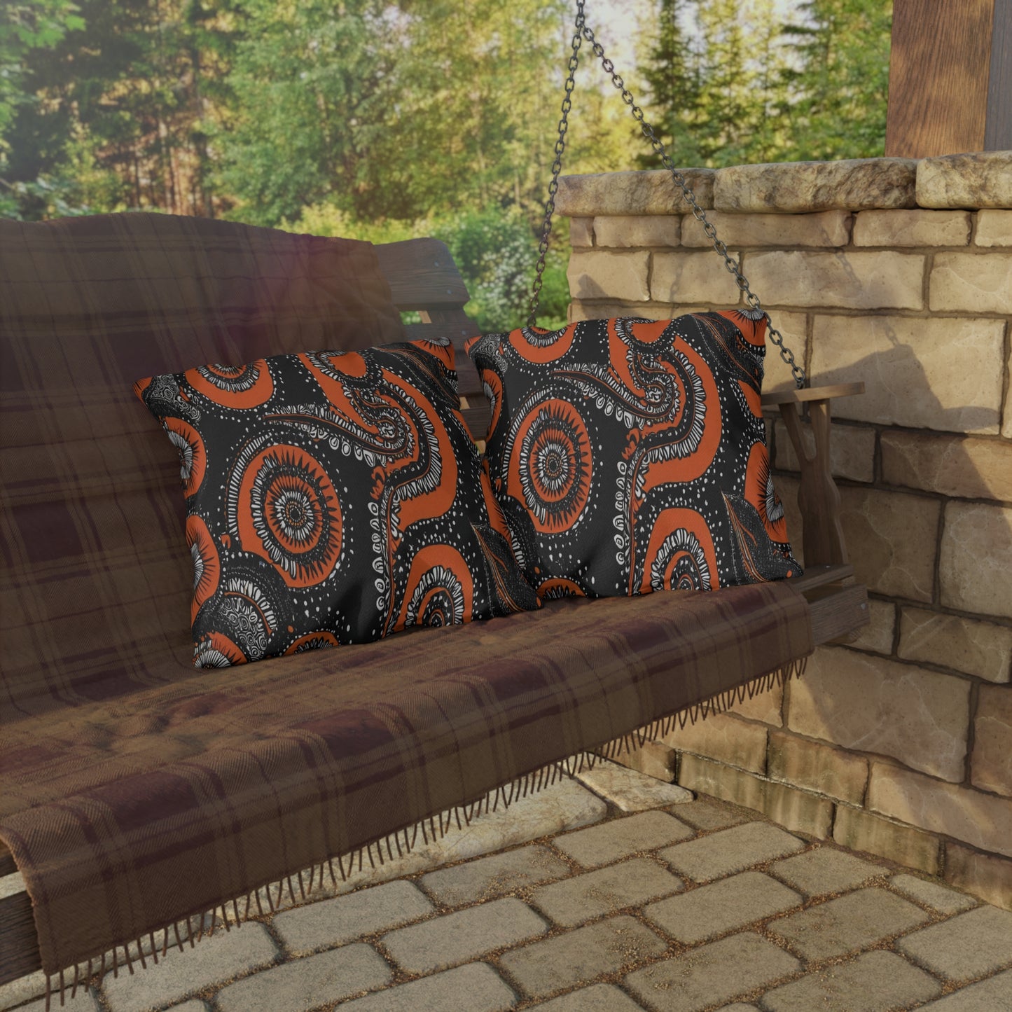 A wooden porch swing with a plaid blanket draped over it holds two African-inspired pillows featuring black, orange, and white abstract circular patterns. The swing hangs beside a stone pillar, with a green forest in the background.
