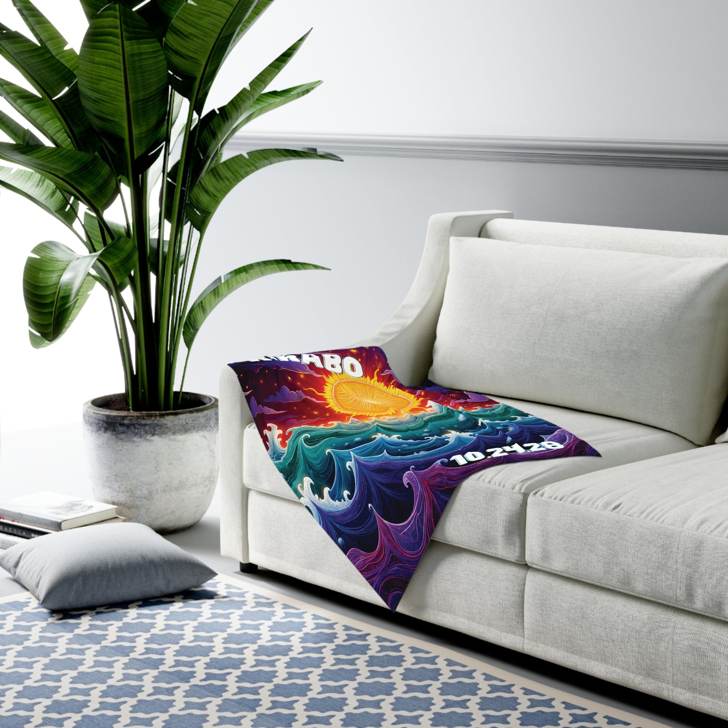 A bright, colorful personalized baby blanket featuring a sun and swirling ocean waves is draped over a modern white sofa. A large potted plant and hints of African-inspired decor complete the stylish, minimal room with a geometric rug and floor pillow.