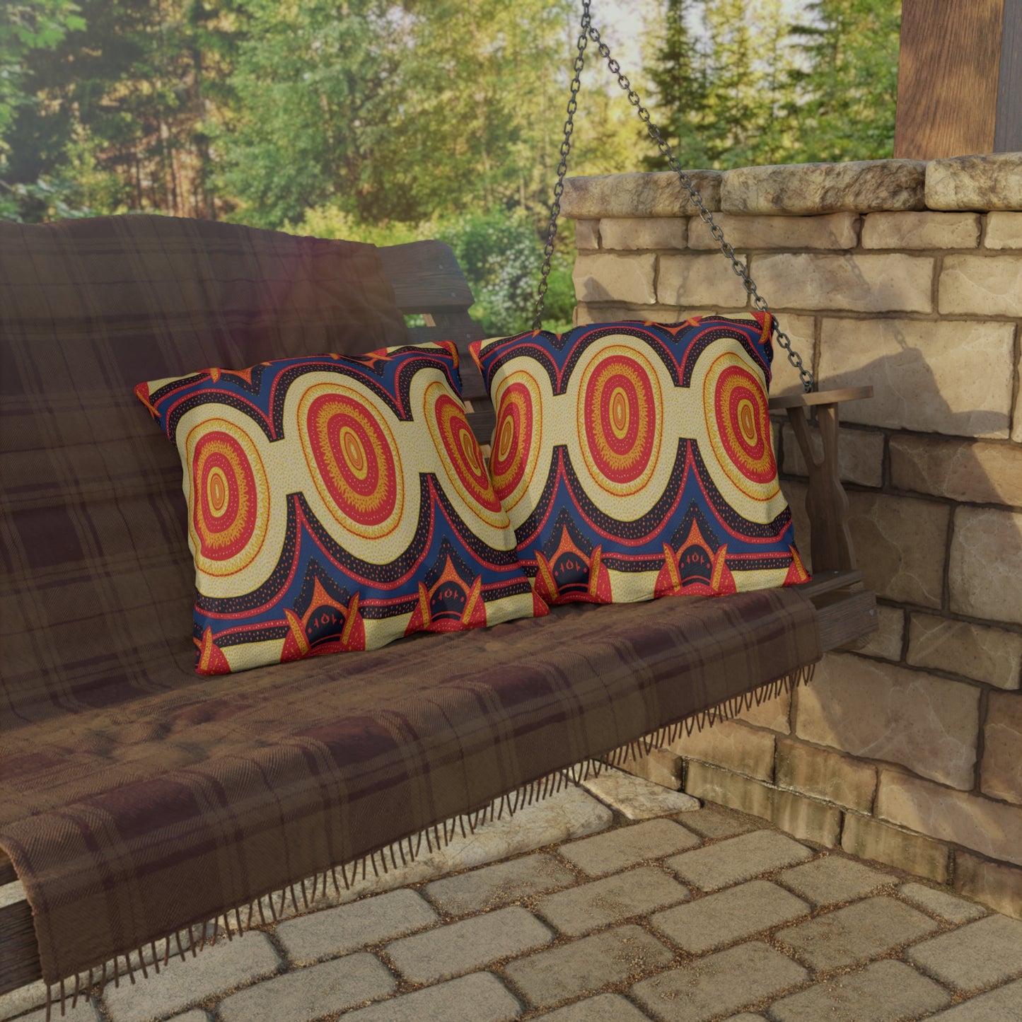 A wooden porch swing covered with a brown plaid blanket holds two colorful, African-inspired pillows featuring bold red, yellow, and blue circular patterns. The swing sits beside a stone wall with sunlit trees in the background.