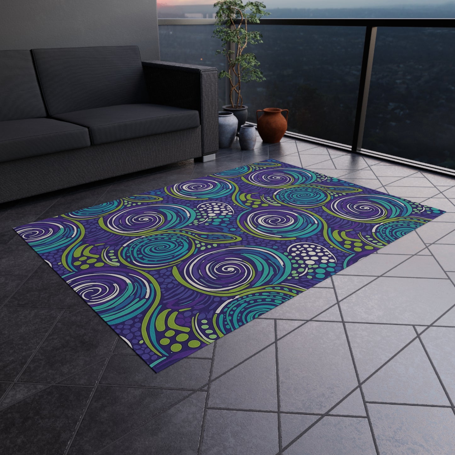 A modern balcony with gray tiled flooring features a colorful, abstract outdoor patio rug in swirling blue, teal, and purple patterns. A dark gray sofa sits against the wall, complemented by potted plants and vases. Cityscape views extend beyond the railing.