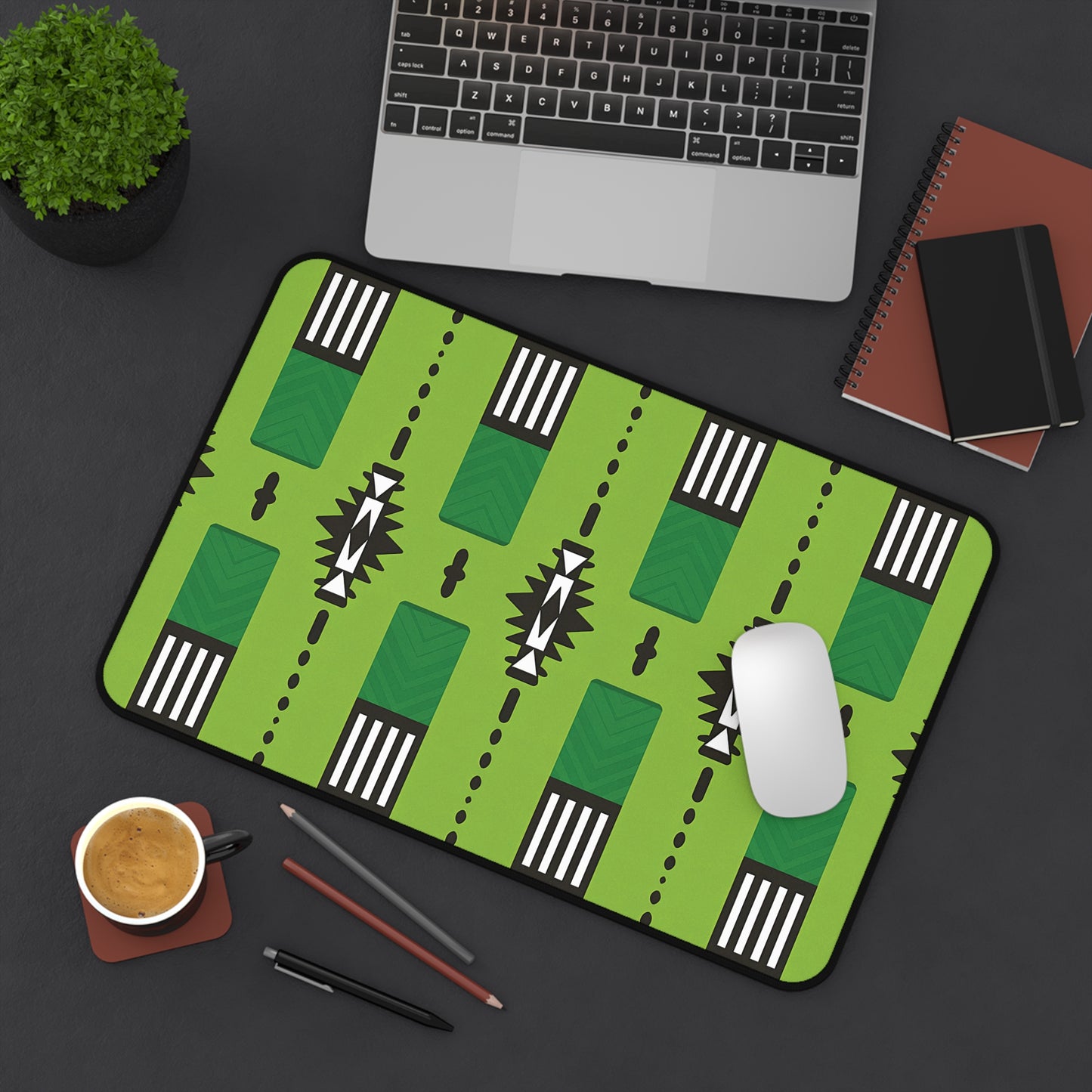 A large green neoprene desk mat with geometric black-and-white designs sits on a dark desk, creating a colorful workspace accessory alongside a laptop, notebooks, smartphone, potted plant, coffee cup, and stationery. A wireless mouse rests on the mat.