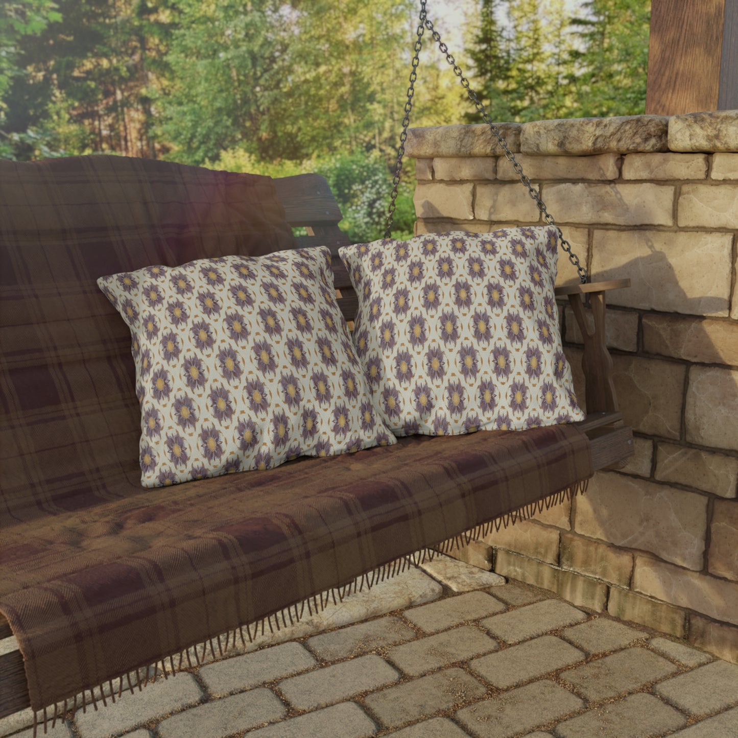 A wooden porch swing with a plaid blanket and two UV-resistant outdoor pillows hangs by metal chains near a stone wall. Sunlight filters through trees in the background, casting shadows on the brick patio below.