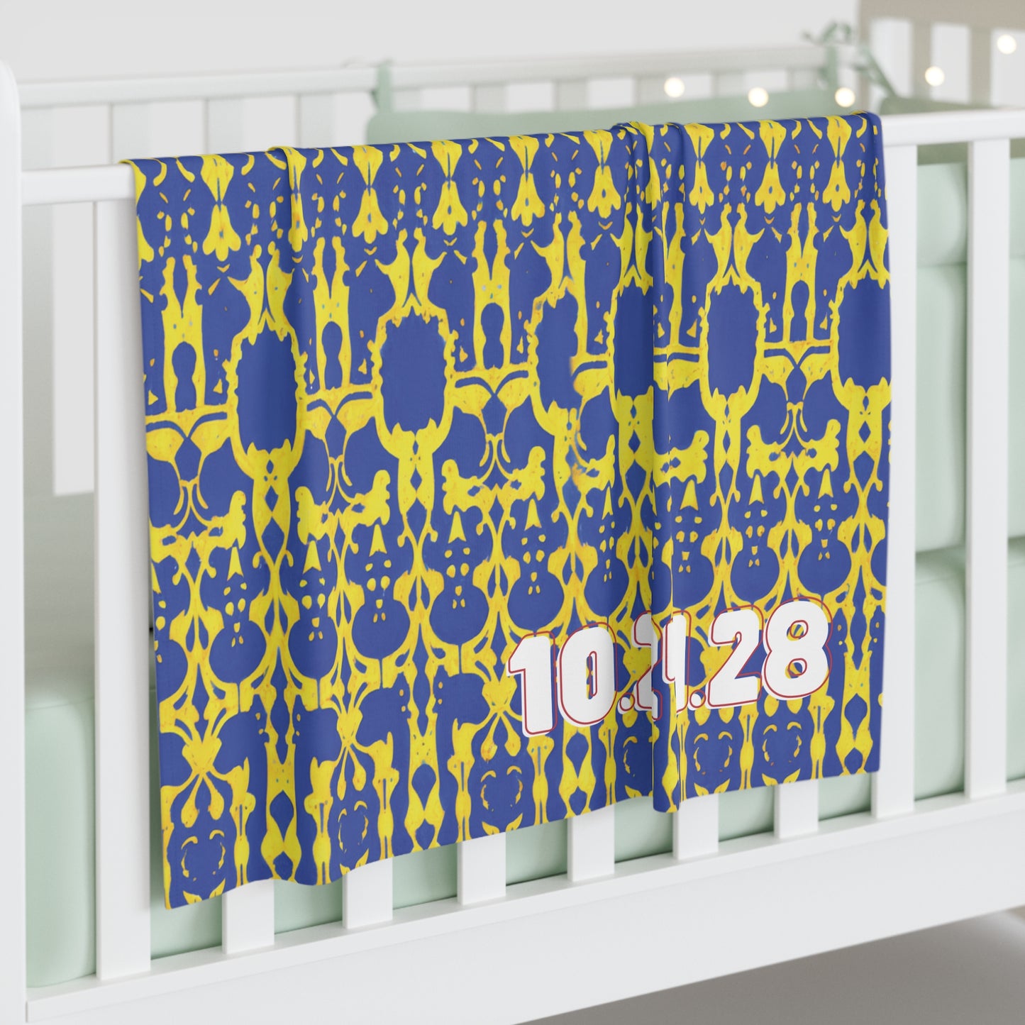 A blue and yellow patterned personalized baby blanket with the numbers 10.1.28 in large, white and pink font drapes over a white crib in a softly lit room, featuring pale bedding and decorative string lights in the background.