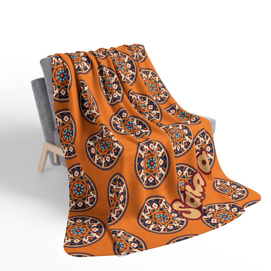 A cozy orange blanket drapes over a gray armchair. The African-inspired design features a repeating pattern of colorful, circular geometric mandalas and a large stylized “Sahara” text in cream and maroon in the bottom right corner.